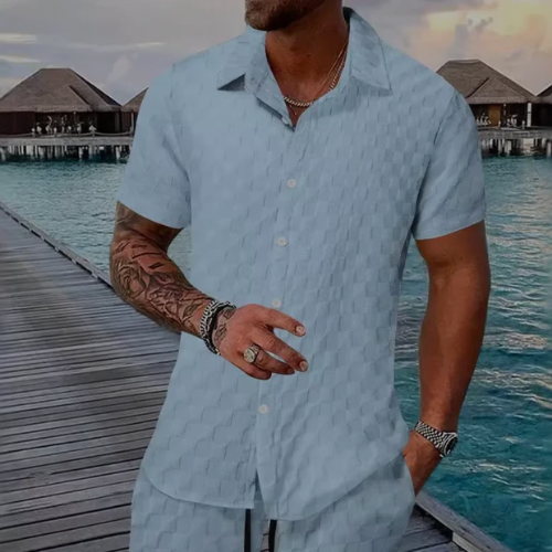Man wearing a light blue patterned shirt by a dock with water and huts in the background