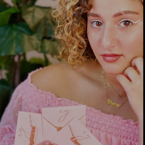 Woman holding jewelry cards with necklaces, wearing a pink top and gold necklace.
